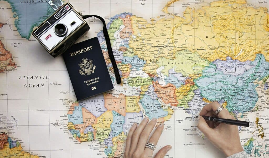 A camera and passport lay on a large map and hands are holding a pen while hovering over a country.