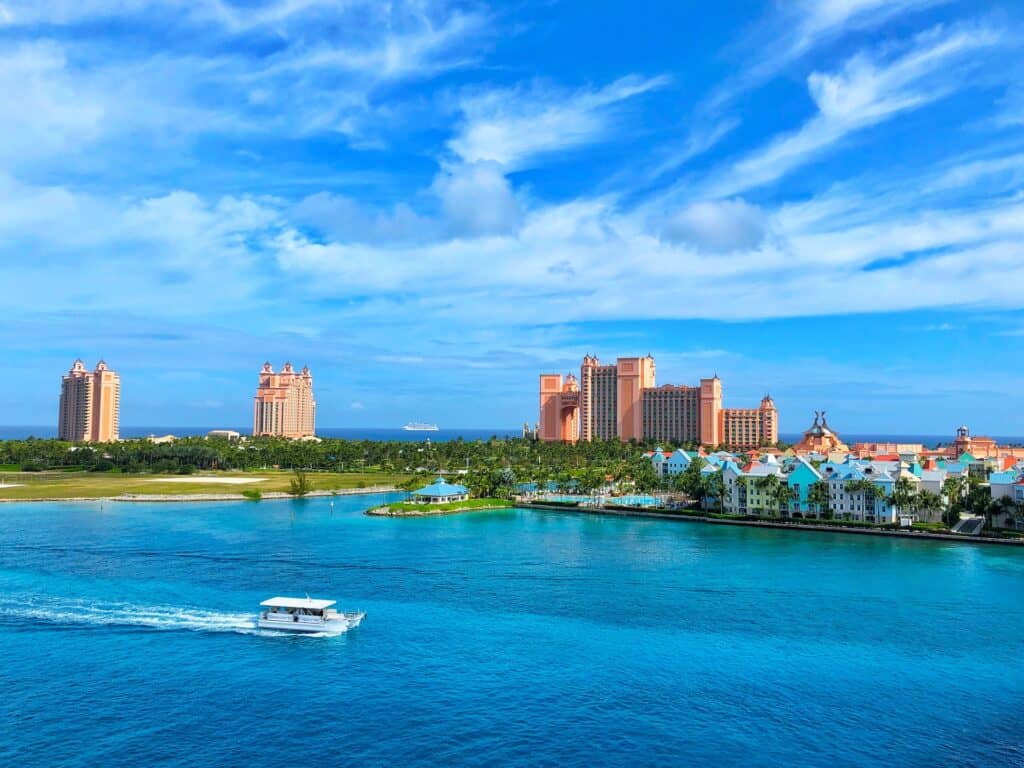 Buildings in the Bahamas nestled among lush greenery along the coastline with a boat cruising on the tranquil water. 
