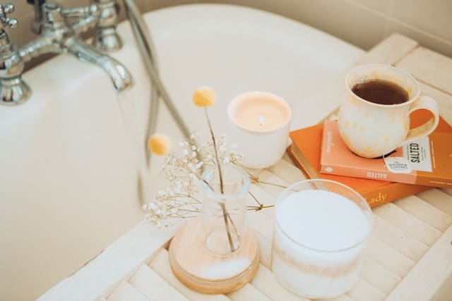 On a wooden tray over a bathtub sits a mug atop books with candles and a small vase with flowers beside it.