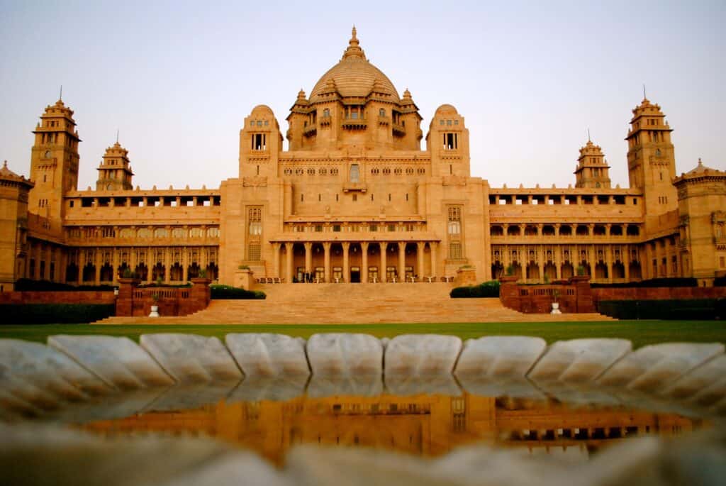 The dusky-tan and grand structure of the Umaid Bhawan Palace sits in front of a sprawling green lawn and unique pond. 