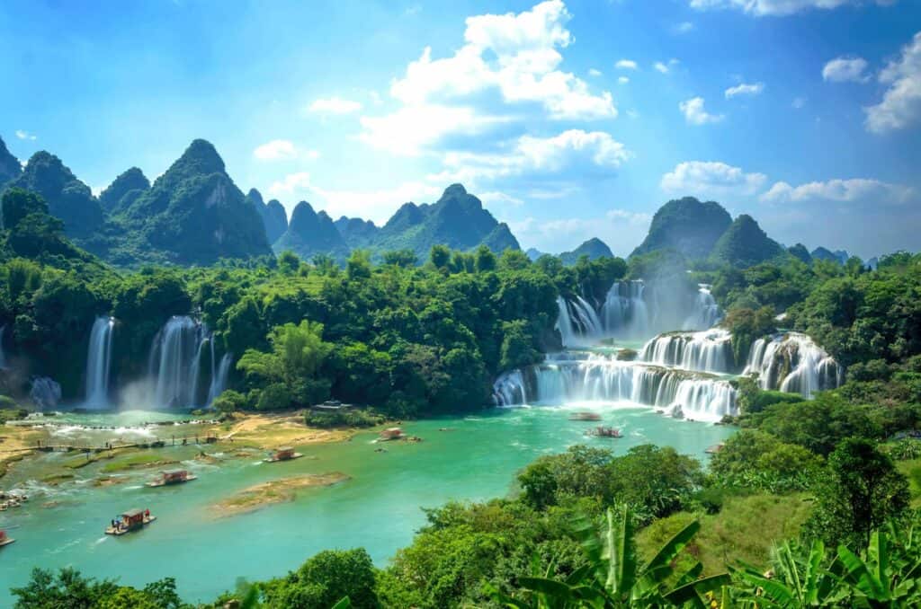Bright sunlight streams over the two sets of waterfalls on the border of China and Vietnam, while raft boats carry visitors to have a closer look at the falls.